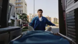 Man in a blue jacket pushing a stretcher from a vehicle onto a city street with buildings in the background.