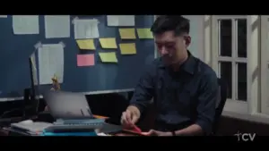 Man working at a desk with a laptop, colorful sticky notes on a wall, focused on a red pen and paper.