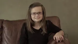 Smiling girl wearing glasses, sitting on a brown leather couch, showcasing a cheerful indoor atmosphere.