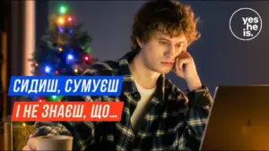 Young man sitting at a laptop with a festive Christmas tree, looking pensive and contemplative in a cozy setting.