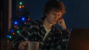 Young man in plaid shirt works on laptop with a festive Christmas tree lit up in the background, holding a coffee cup.