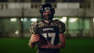 Football player in camouflage uniform holds a ball, standing on a field at night with stadium lights in the background.