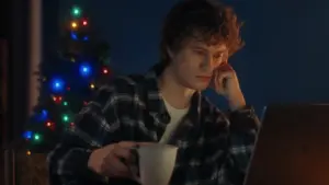 Person in flannel shirt sitting with a mug, gazing thoughtfully at a laptop, festive tree lights in the background.