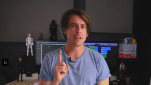 Young man in a blue shirt gestures thoughtfully, with computer screens and figurines visible in a modern workspace.