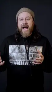 Man with long hair and beard wearing a graphic hoodie, gesturing passionately against a dark background.