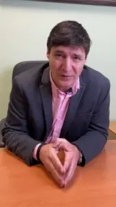 Man in a gray blazer and pink shirt sitting at a desk, gesturing with hands during a discussion.