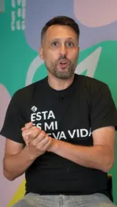 Man in a black t-shirt expressing enthusiasm while seated against a colorful background.