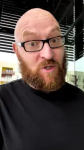 Man with a bald head and red beard wearing glasses, making a surprised expression in a modern indoor setting.