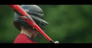 Young baseball player preparing to hit, wearing a black helmet and holding a red bat, set against a green background.