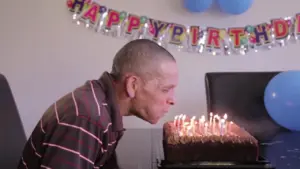 A person blowing out candles on a birthday cake, with a "Happy Birthday" banner and blue decorations in the background.