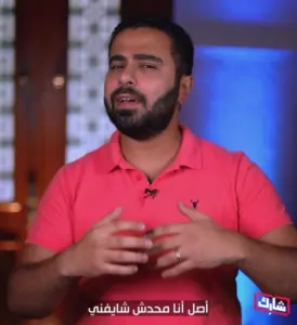 Man in a pink shirt gesturing while speaking, with a blurred background and Arabic subtitles displayed.