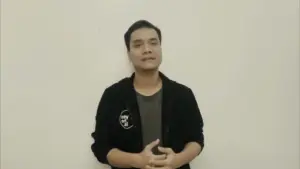 Young man in a black jacket standing against a plain white wall, speaking confidently with hands clasped.