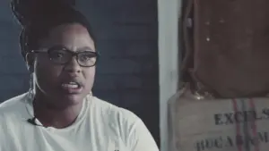 Woman with glasses speaking passionately in a dimly lit room, conveying a strong message against a textured background.