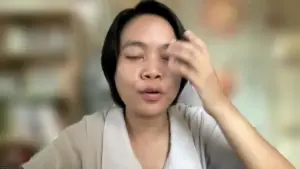 A woman in a light-colored top gestures thoughtfully while touching her forehead during a virtual meeting.