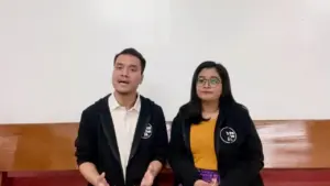 Two presenters in black jackets speaking on stage against a plain white background, engaging the audience.