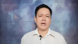 A man with short black hair speaks against a colorful, abstract background, wearing a white collared shirt.