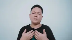 Man in a black shirt gesturing with both hands against a light-colored background, expressing a thoughtful demeanor.