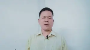 Man speaking against a plain white background, wearing a light yellow shirt and a microphone attached to his collar.