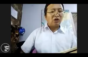 Person speaking during a virtual meeting, wearing glasses and a white shirt, in an office setting with a map in the background.