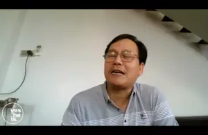 Man in a striped shirt sits indoors, smiling while speaking, with a staircase visible in the background.