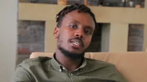 Man with short, twisted hair sitting on a couch, engaging in conversation with a thoughtful expression.