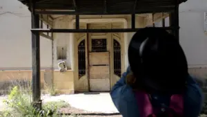 A person with a black hat stands in front of an old, abandoned building entrance with peeling paint and a deserted path.