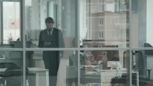 Businessman in a suit standing alone in a modern office, looking at his phone through glass windows.