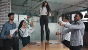 Team celebrating success in a modern office, with one member joyfully standing on a table surrounded by colleagues.