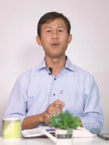 Person sitting at a table with a book, engaging in spoken presentation, complemented by a small green plant and a mug.