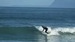 Surfer riding a wave at a scenic beach with distant mountains under a clear blue sky.