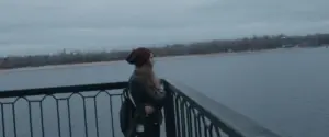 Person in a beanie gazing over a calm lake, with a cloudy sky and scenic landscape in the background.