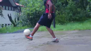 Person dribbling a soccer ball on a wet surface, surrounded by lush greenery and a rustic building in the background.