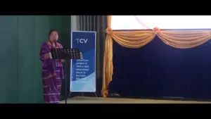 Speaker presenting at an event, wearing traditional attire, with a backdrop featuring a blue banner and golden drapes.