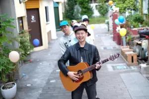 A musician playing guitar in a vibrant alley, surrounded by colorful balloons and smiling friends.