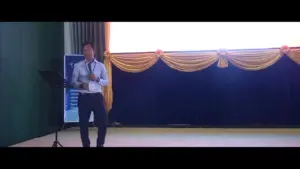 Presenter on stage speaking at a conference, with a backdrop of decorative drapes and a presentation screen.