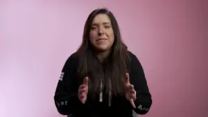 Young woman in a black hoodie sitting against a pink background, gesturing with her hands while speaking.