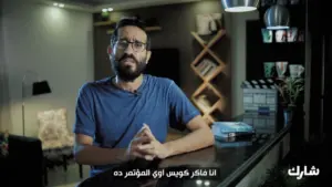 Man speaking at a desk in a modern room, discussing a topic with a thoughtful expression and cinematic elements visible.