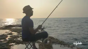 A person fishing at sunset, seated on a stool by the shore, with a serene ocean backdrop and a fishing rod in hand.