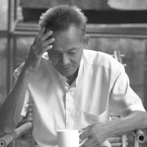 Elderly man in a white shirt thoughtful while holding a white cup, captured in a serene black and white setting.