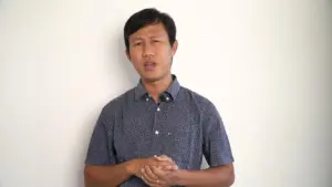 Man standing against a white wall, looking thoughtfully with hands clasped, wearing a blue patterned shirt.