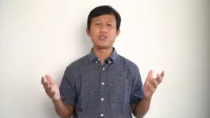 Man in a patterned shirt standing against a white wall, gesturing with hands to emphasize a point.