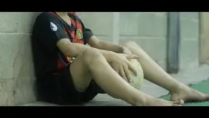 Child in a red and black sports jersey sitting against a wall, holding a rugby ball with bare feet.