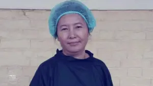 Smiling woman in surgical cap and gown stands against a beige brick wall, representing healthcare and professionalism.