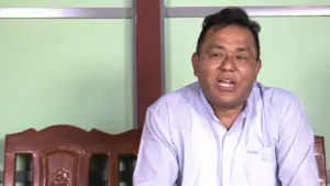 A man in a light blue shirt sits on a wooden chair, speaking confidently against a simple green backdrop.