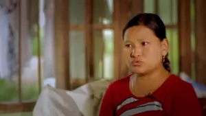 Woman in a red shirt sitting indoors, expressing emotions with a thoughtful gaze, natural light illuminating the scene.