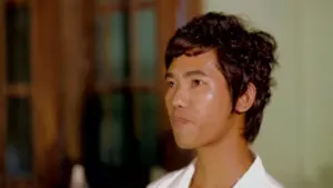 Young man with curly hair in a white shirt, smiling slightly, set against a softly lit indoor background.