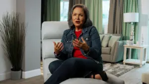 Woman in denim jacket sitting on a chair, gesturing while speaking in a cozy, well-lit living room setting.