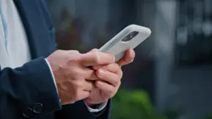 Person in a suit holding a smartphone, focusing on the device while outdoors in a blurred green background.