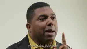 Man in a yellow shirt passionately speaking, gesturing with one finger raised, against a soft neutral backdrop.