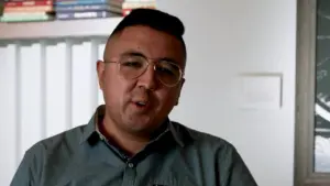 Man with glasses speaking indoors, surrounded by shelves of books and modern decor. Engaging and informative expression.